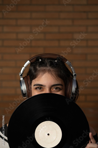 Mujer joven de pelo negro caucásica con auriculares y un disco de vinilo negro tapando su cara, enseñando sus ojos, sonriendo