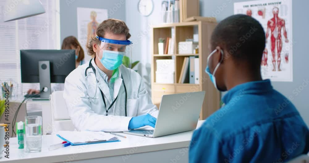 Portrait of Caucasian male general practitioner sitting in medical mask and protective face shield in hospital office with African American male patient writing down symptoms and typing on laptop Portrait of Caucasian male general practitioner sitting in medical mask and protective face shield in hospital office with African American male patient writing down symptoms and typing on laptop