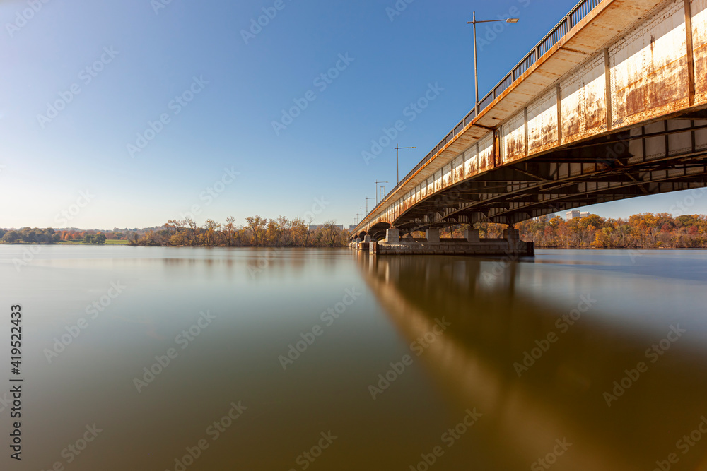 Long exposure landscape image featuring Theodore Roosevelt Island and bridge and Potomac river on a sunny afternoon in autumn. Trees on island have orange red brown colors. The bridge is rusty .