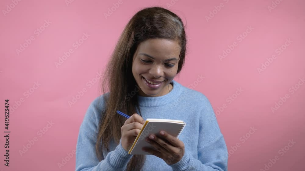 African woman making notes in planner, girl holding pen. She writes future plans and to-do list in notebook for week or month. Keeping personal diary on pink studio background. African woman making notes in planner, girl holding pen. She writes future plans and to-do list in notebook for week or month. Keeping personal diary on pink studio background.