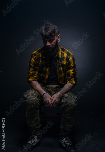 Handsome male model with tattoos and tartan shirt is sitting on a chair isolated on a black background