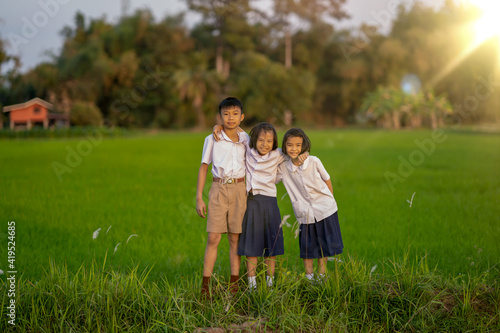 Familly child wearing students uniform playing on rice field background on sunset, cute Asian kid smiling and holding flower in countryside, child standing on grass field of rice agriculture view
