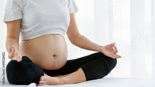 close up of a tummy of a healthy pregnant woman sitting on a white bed doing Yoga for excise in a bedroom at home. Selective focus on the belly of a lady