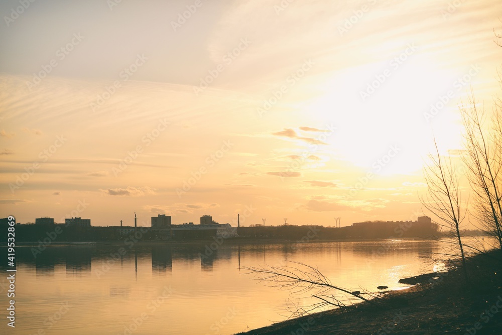 Scenic view of a beautiful sunset over the river in the city in spring against the background of the golden sky, the sun over the water and silhouettes of bushes in the foreground. Spring landscape.
