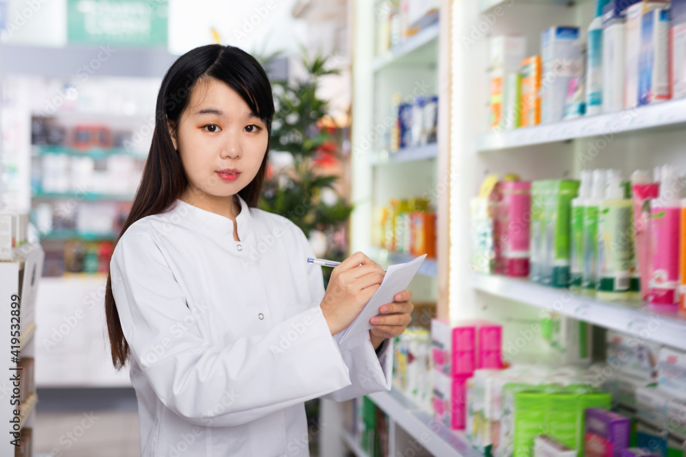 Glad cheerful positive smiling chinese woman pharmacist keeps track of drugs in interior of pharmacy