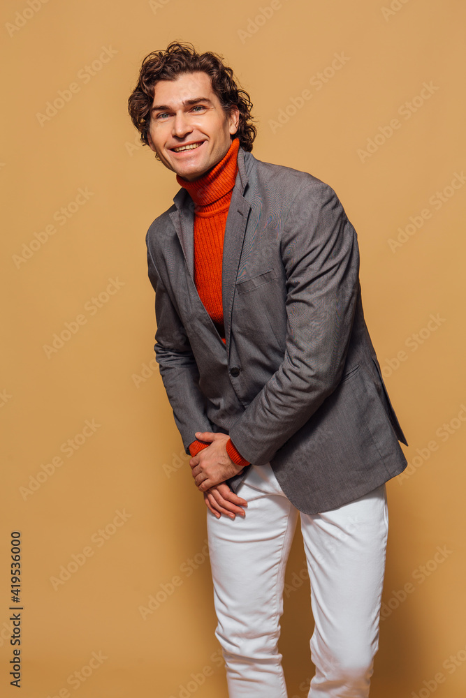 Obraz premium Tall handsome man dressed in orange turtleneck, white jeans and grey jacket posing on the beige background
