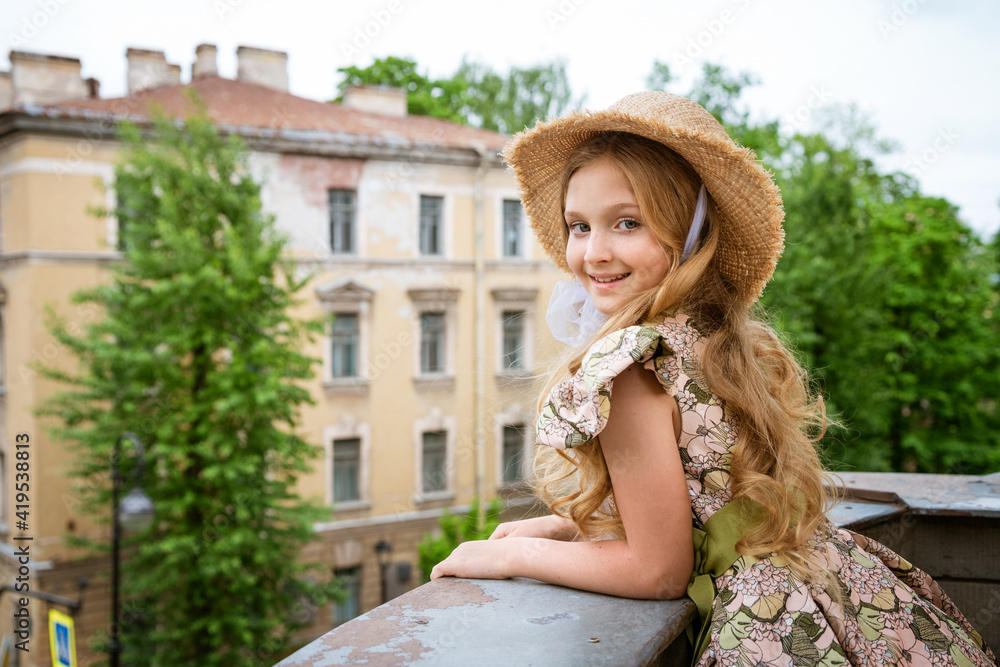 Little beautiful girl in dress and hat posing on the balcony against the background of the city