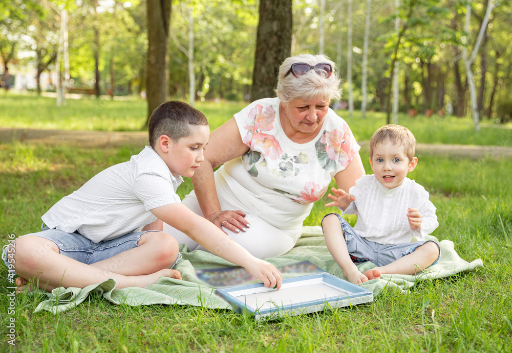 Fototapeta premium Cheerful extended family playing board game in their backyard. Family playing in the summer forest. Grandmother playing with her grandchildren at nature. Happy moments with grandmother outdoors.