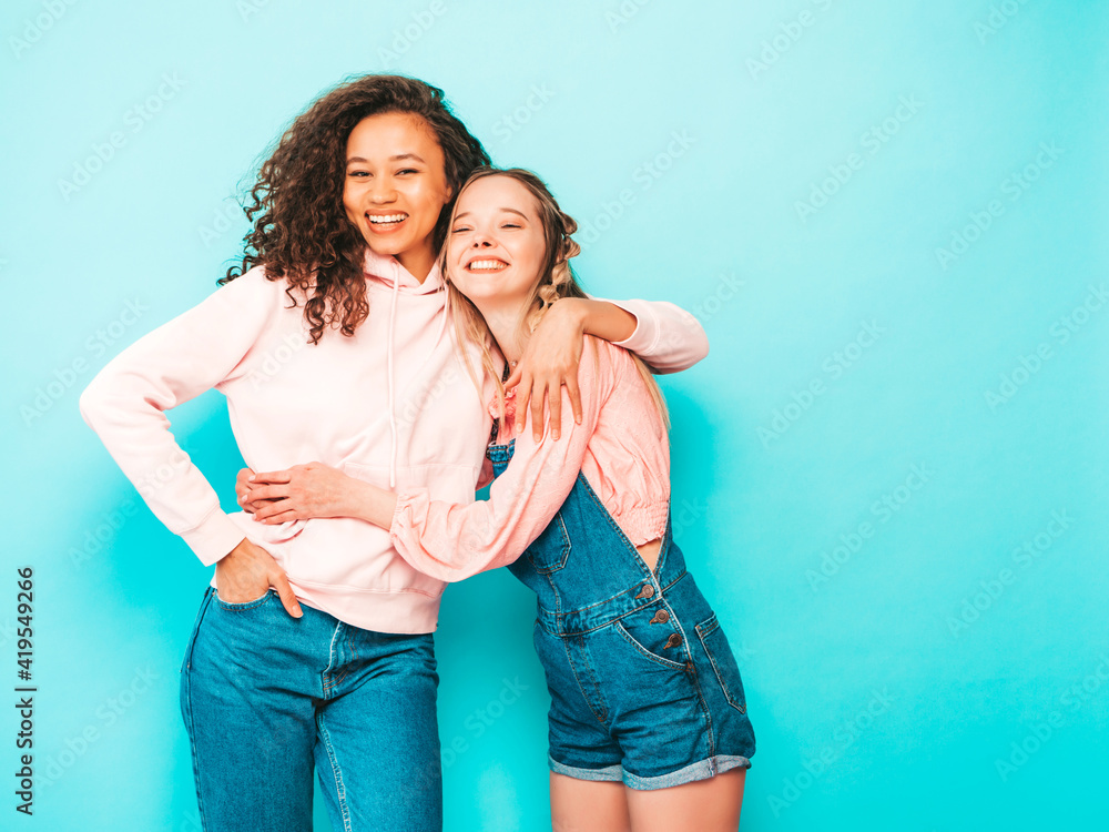 Two young beautiful smiling international hipster female in trendy summer clothes. Sexy carefree women posing near blue wall in studio. Positive models having fun in sunglasses. Concept of friendship Two young beautiful smiling international hipster female in trendy summer clothes. Sexy carefree women posing near blue wall in studio. Positive models having fun in sunglasses. Concept of friendship