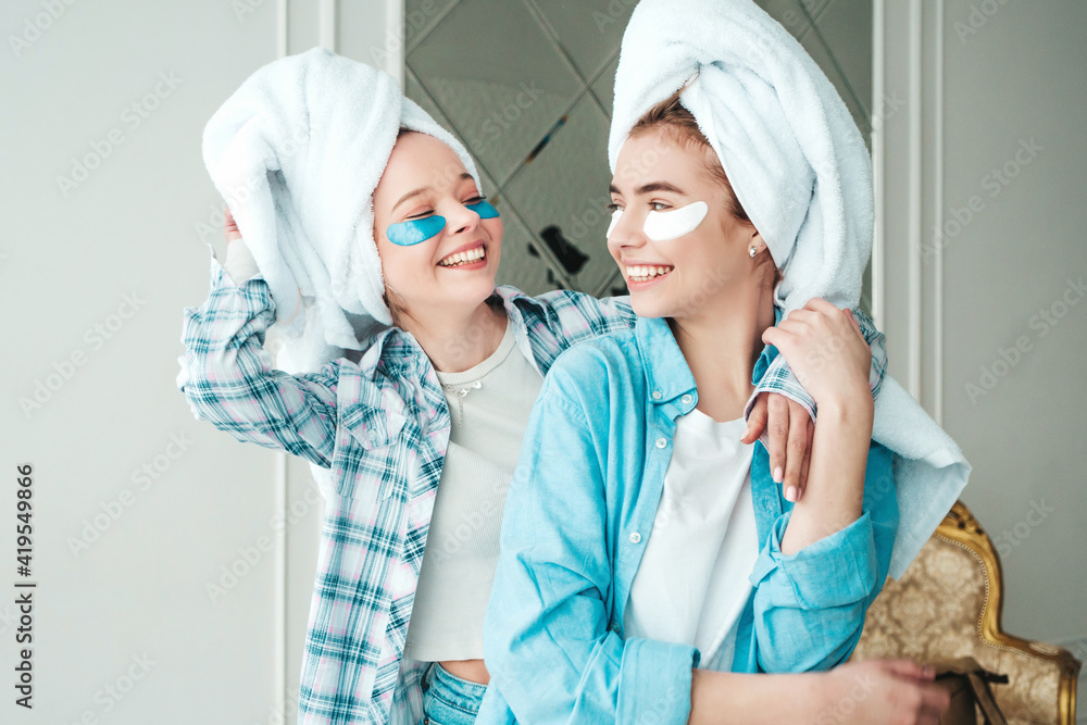 Two young beautiful smiling women with patches under their eyes. Sexy carefree models sitting on bed in posh apartment or hotel room. They doing beauty treatments at home in towels on heads Two young beautiful smiling women with patches under their eyes. Sexy carefree models sitting on bed in posh apartment or hotel room. They doing beauty treatments at home in towels on heads