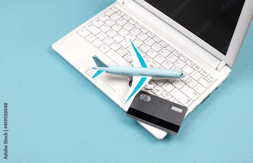 White laptop on a blue background. Nearby is a bank card and a miniature airplane. Concept - Online booking of air tickets, travel planning. Payment for orders via the Internet. White laptop on a blue background. Nearby is a bank card and a miniature airplane. Concept - Online booking of air tickets, travel planning. Payment for orders via the Internet.