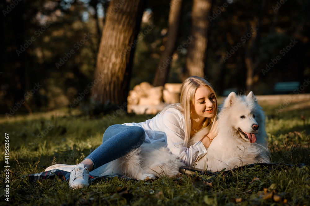 beautiful woman in white shirt is hugging her white dog samoyed outdoors in the park and sitting on the grass. beautiful woman in white shirt is hugging her white dog samoyed outdoors in the park and sitting on the grass.