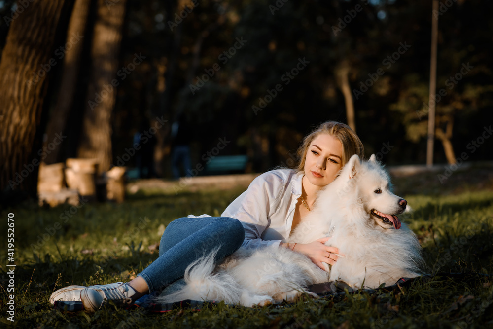 beautiful woman in white shirt is hugging her white dog samoyed outdoors in the park and sitting on the grass. beautiful woman in white shirt is hugging her white dog samoyed outdoors in the park and sitting on the grass.