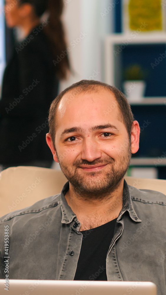 Manager sitting on couch in front of camera smiling while multiethnic team working in background with financial reports. Diverse coworkers talking about startup financial company in modern office. Manager sitting on couch in front of camera smiling while multiethnic team working in background with financial reports. Diverse coworkers talking about startup financial company in modern office.