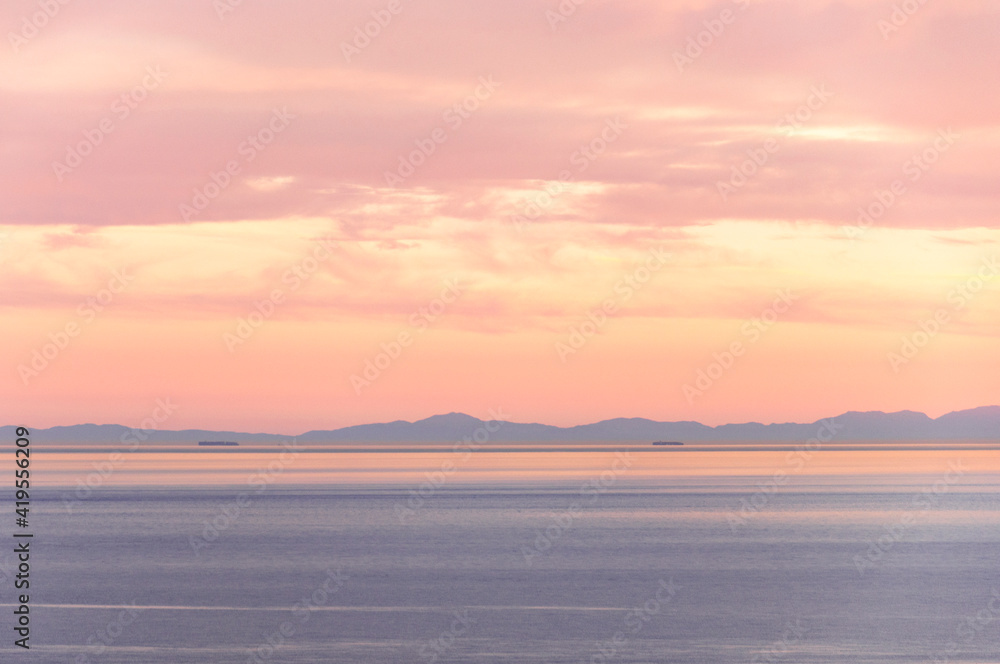 Pastel shades of pink and blue colors, a rippled sea with two ships in the distance, the opposite shore and a sky with clouds in the first seconds after sunset.