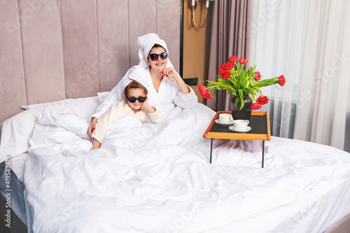Happy beautiful family mother and daughter in the bedroom on the bed in towels and sunglasses arranged a fashionable party