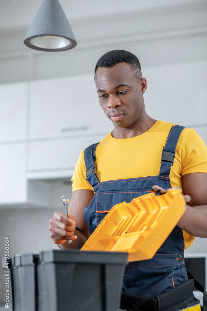 Dark-skinned service man standing in the kitchen and opening his tools box Dark-skinned service man standing in the kitchen and opening his tools box