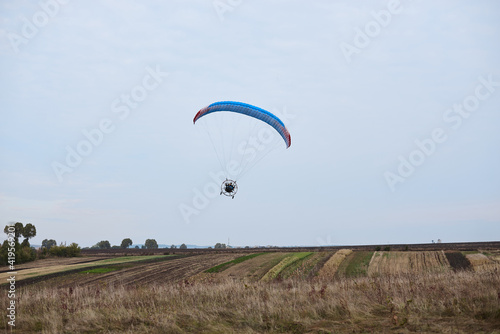 Paratrike with blue parachute flying high in the cloudy sky on autumn morning. Paragliding activity in rural countryside. Weekend leisure time and hobby. Extreme sport flight on paramotor machine.