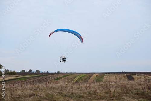 Paratrike with blue parachute flying high in the cloudy sky on autumn morning. Paragliding activity in rural countryside. Weekend leisure time and hobby. Extreme sport flight on paramotor machine.