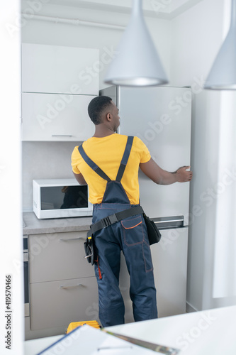 Dark-skinned service man in yellow tshirt repairing the fridge