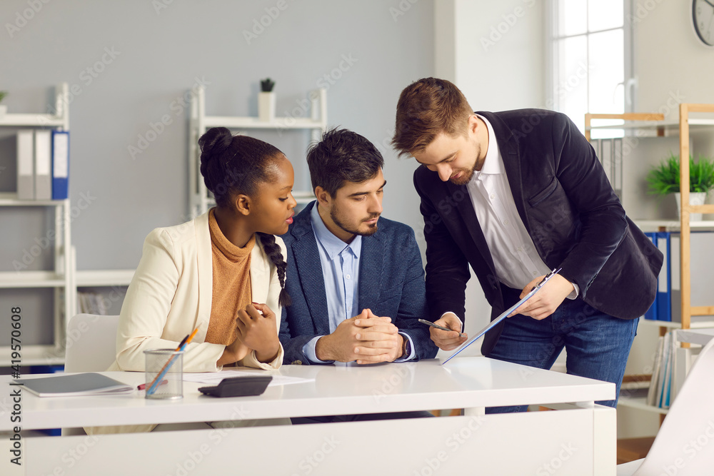 Bank employee gives documents for signature to a Caucasian man and his dark-skinned wife. Specialist in lending and financial assistance for young families indicates the place of signature. Bank employee gives documents for signature to a Caucasian man and his dark-skinned wife. Specialist in lending and financial assistance for young families indicates the place of signature.