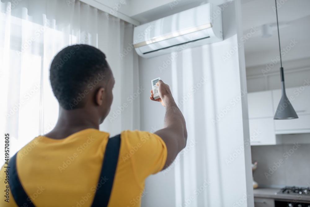 Fototapeta premium Dark-skinned service man in yellow tshirt switching on the air-conditioner