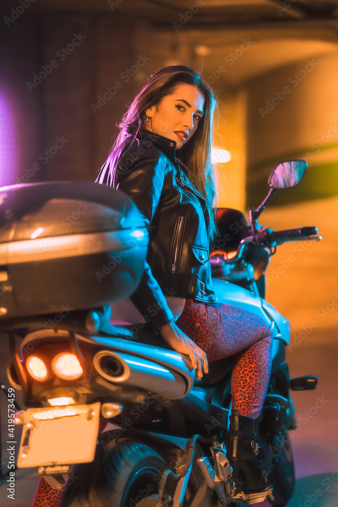 Photography with blue and pink neons on a motorcycle from behind. Portrait of a young blonde Caucasian model wearing a black leather jacket