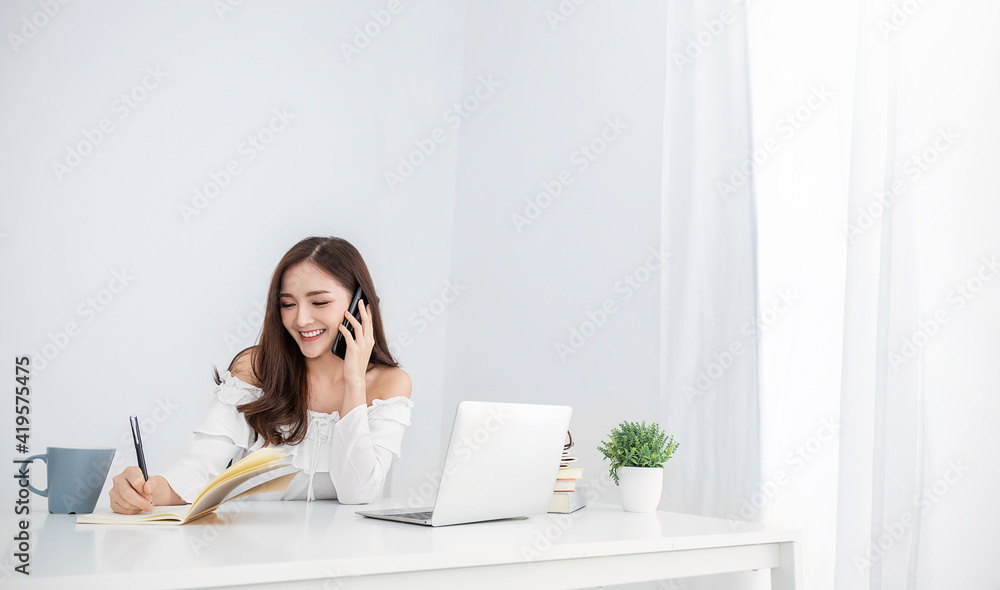 Portrait of young asian business woman talk on smartphone write notebook laptop in home office. Beautiful girl at desk computer take home order. Startup business asia woman online sme telemarketing. Portrait of young asian business woman talk on smartphone write notebook laptop in home office. Beautiful girl at desk computer take home order. Startup business asia woman online sme telemarketing.