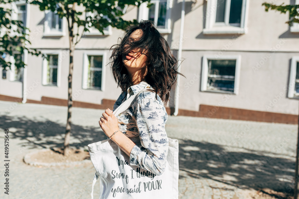 Candid portrait of a joyful young woman walking to the grocery with an eco bag in the street. Positive girl posing with a reusable textile bag on a sunny day.
