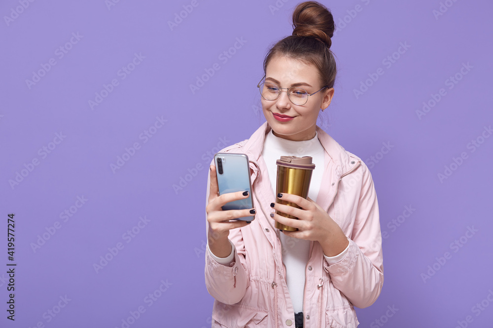 Pleasant looking female wearing spectacles and pale rose jacket posing isolated over lilac background, drinking hot beverage from thermo mug, holding smart phone in hands and browsing Internet. Pleasant looking female wearing spectacles and pale rose jacket posing isolated over lilac background, drinking hot beverage from thermo mug, holding smart phone in hands and browsing Internet.