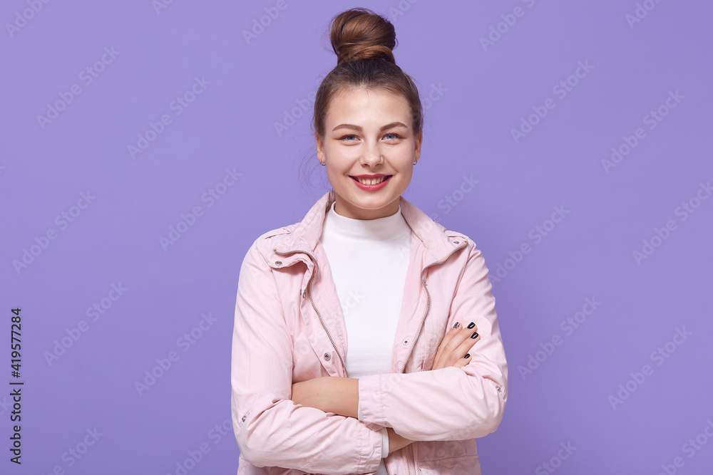 Smiling young woman girl in pale pink jacket and white shirt posing isolated on lilac background, keeping hands crossed, female with hair bun looks at camera, expressing positive emotions. Smiling young woman girl in pale pink jacket and white shirt posing isolated on lilac background, keeping hands crossed, female with hair bun looks at camera, expressing positive emotions.