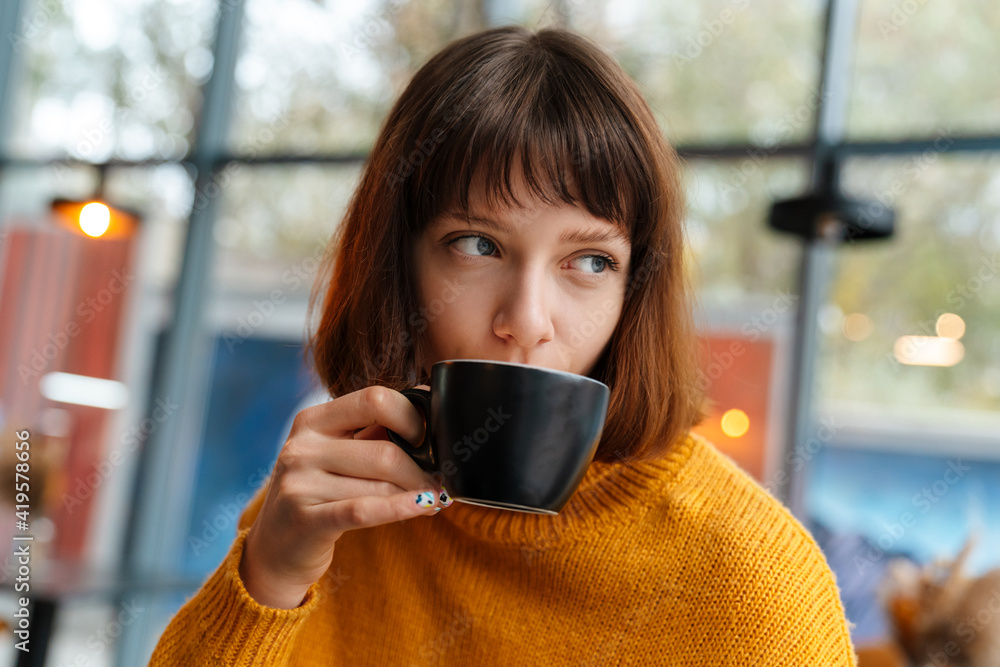Beautiful redhead girl drinking coffee while sitting in cafe indoors Beautiful redhead girl drinking coffee while sitting in cafe indoors
