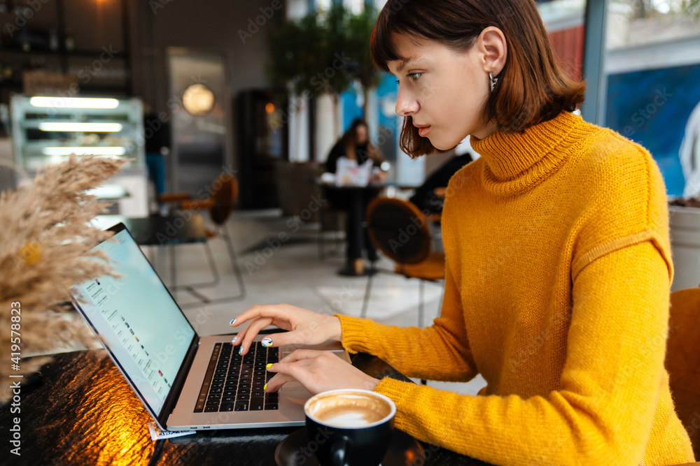 Focused redhead girl drinking coffee and working with laptop in cafe Focused redhead girl drinking coffee and working with laptop in cafe