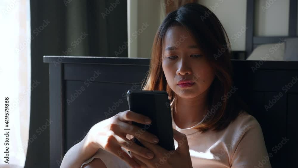 4K Depressed Asian woman sitting by the window in bedroom using mobile phone with boredom. Loneliness sad female being alone with stressed and negative emotion. Mental health and life problem concept. 4K Depressed Asian woman sitting by the window in bedroom using mobile phone with boredom. Loneliness sad female being alone with stressed and negative emotion. Mental health and life problem concept.