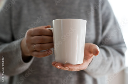 Close-up of female hands with a mug of drink. A beautiful girl in a gray sweater holds a cup of tea or coffee in the morning sunlight. Mug for your design. Empty space.