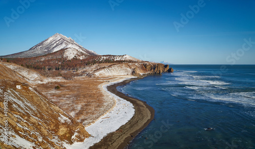 Deep blue sea with foamy waves has winding borders with the sand coast in winter. A field with dry grass and hills around.