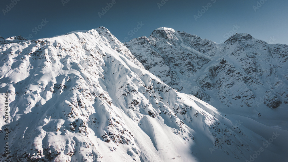 Amazing old mountains with steep slopes covered with shiny snow and shadows under blue sky in sunny winter morning aerial view