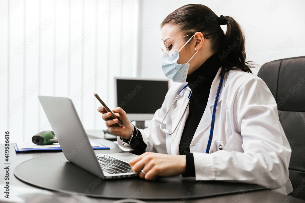 Young doctor working on laptop in office analyzes medical records of his patients infected whit Coronavirus Covid-19 wearing protective face mask take a break looking a message on smartphone Young doctor working on laptop in office analyzes medical records of his patients infected whit Coronavirus Covid-19 wearing protective face mask take a break looking a message on smartphone