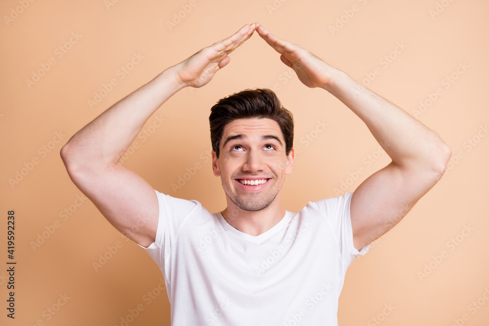Photo of cheerful guy look up empty space arms show house shape toothy smile isolated on beige color background Photo of cheerful guy look up empty space arms show house shape toothy smile isolated on beige color background