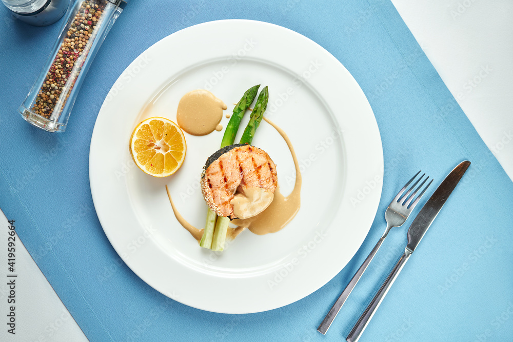 Appetizing salmon steak with asparagus and sauce in a white plate on a blue tablecloth.