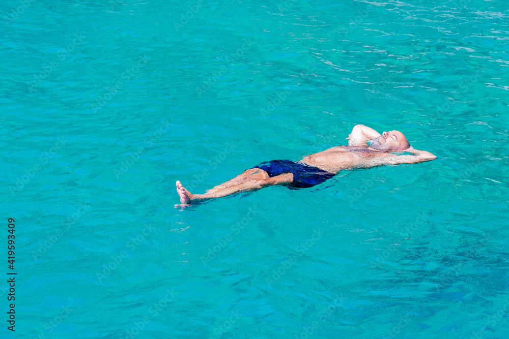 Summer concept: A single Caucasian man with blue shorts is lying on the water. The guy is floating fearless over turquoise sea. Outdoors on a sunny day. Swimming activity on holiday.