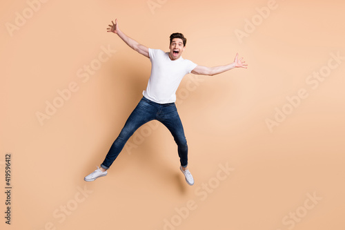 Full size photo of optimistic brunet man jump yell wear t-shirt jeans sneakers isolated on beige color background