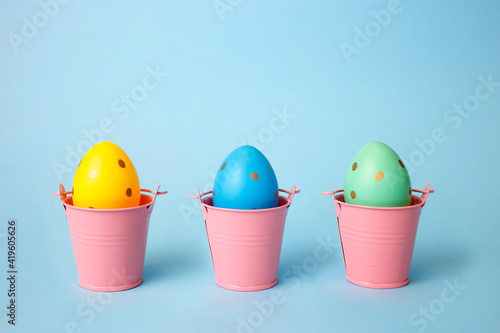 Three colored decorated eggs in buckets on a blue background. Easter greeting card in minimalist style concept.Copy space