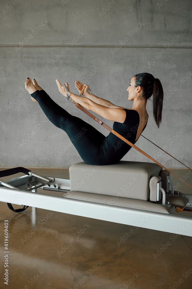 Foto de Full shot of a strong body of a Pilates Instructor in V sit on a  bed of a Reformer machine, pulling a stretch band with hands, training in a  big