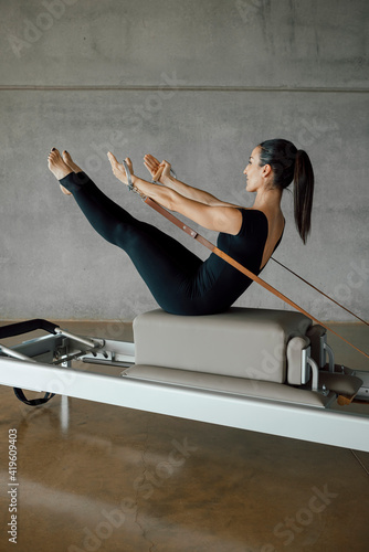 Full shot of a strong body of a Pilates Instructor in V sit on a bed of a Reformer machine, pulling a stretch band with hands, training in a big Industrial Fitness Center Studio. Full length.