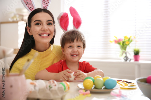 Happy mother with her cute son painting Easter eggs at table indoors