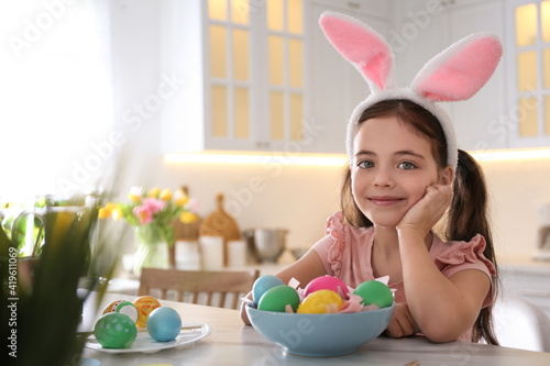 Cute little girl in bunny ears headband painting Easter eggs at table indoors