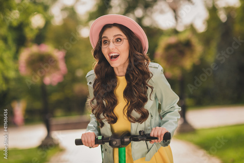 Photo of impressed excited young woman dressed teal jacket glasses cap riding kick scooter outdoors city park