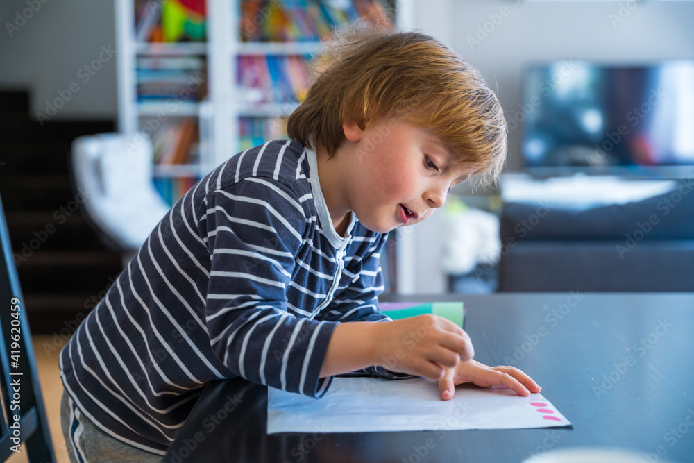 Little boy sitting at the table, learning at home during coronavirus quarantine. Toddler looking at the early development book. Child educating in playing with stickers. Little boy sitting at the table, learning at home during coronavirus quarantine. Toddler looking at the early development book. Child educating in playing with stickers.