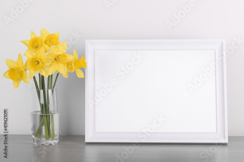 Breakfast still life., empty picture frame mockup on wooden desk, table. Vase with daffodils branches. Elegant working space, home office concept. Scandinavian interior design